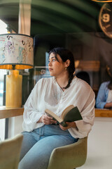 Chica joven guapa en cafetería sentada junto a una ventana con un libro 