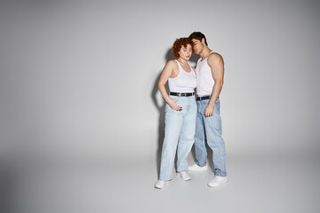 appealing sexy couple in blue jeans posing together lovingly on gray background, relationship