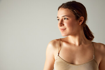 Portrait of beautiful smiling young girl with brunette hair in ponytail, posing in underwear against grey studio background. Concept of body and health care, female beauty, wellness