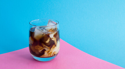 Iced coffee with milk in the drinking glass on the pink and blue background. Close-up.