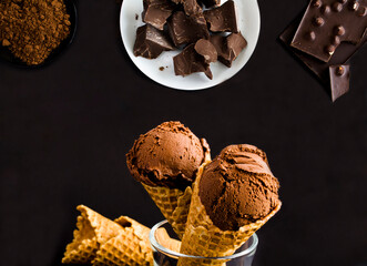 Chocolate ice cream in a waffle cone, chunks of dark chocolate and cocoa powder on the black background. Close-up.