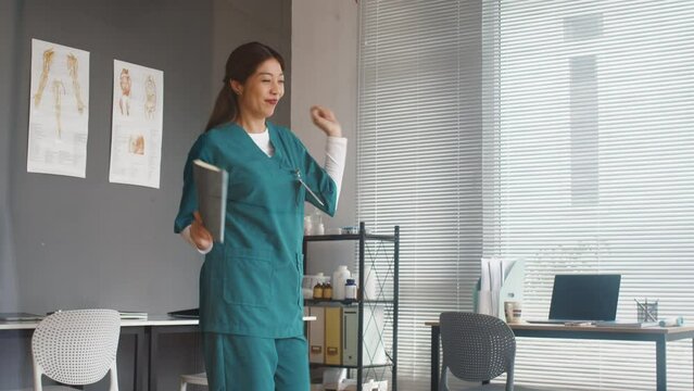 Medium long shot of cheerful Asian medical worker in uniform dancing in nurses room after passed exam in clinic
