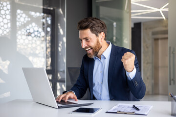 Fototapeta premium Happy young businessman sitting in the office at the table and looking at the laptop screen, rejoices at the success, received information, showing a victory gesture with his hand