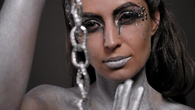 Close-up of a young woman with silver paint on her skin holding a silver chain and posing in front of the camera in the studio. Portrait of a fashion woman with silver body art.