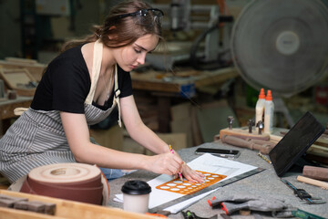 A young woman is training to be a carpenter in the workshop. She works with a laptop computer in a wood workshop. female carpenter contact customers by smartphone. SME
