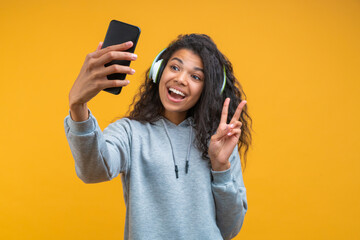 Studio portrait of cute happy smiling girl taking selfie on her mobile phone while listening to music via wireless headphones