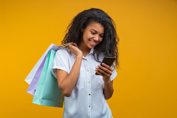 Studio portrait of beautiful young woman with charming smile standing with shopping bags in hand while using her smart phone, isolated over bright colored yellow background