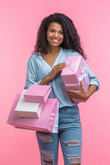 Studio vertical portrait of happy smiling beautiful casually dressed young woman posing standing with gift box and bunch of shopping bags in hands over pastel pink background