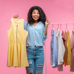 Studio portrait of excited smiling attractive young woman holding hanger with illuminating yellow dress in hand while standing near the rack of pastel clothes in showroom