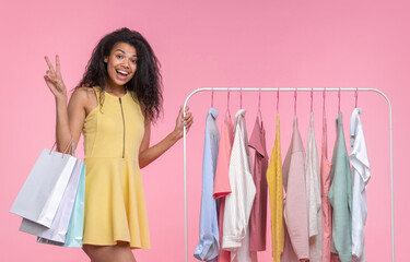 Studio portrait of cute and funny girl posing over pastel pink background with a bunch of shopping bags, showing v-sign gesture while standing near clothing rack