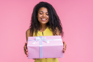 Studio shot of smiling beautiful birthday girl looking at the decorated present box in her hands with content face expression, isolatd over pastel pink background - 745787109