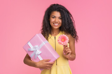 Studio shot of beautiful smiling woman in pastel yellow dress holding decorated gift box and gerbera flower in hand