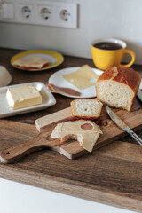 a bitten sandwich on a wooden board, next to a yellow cup of tea, butter in a butter dish, cheese and meat on a plate, a knife, a kitchen towel and a vase of flowers. breakfast in a white kitchen.