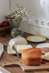 sliced bread on a wooden board, next to butter in a butter dish, cheese and meat on a plate, a knife, a kitchen towel and a vase with gypsophila flowers in a white kitchen. breakfast in the kitchen