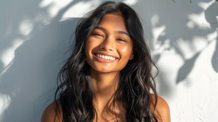 Smiling woman with long dark hair and glowing skin standing in soft light with a white wall and tree shadows behind her. - obrazy, fototapety, plakaty