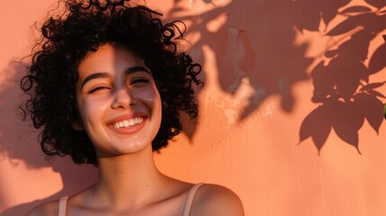 A joyful young woman with curly hair smiling brightly against a warm orange wall with shadows of leaves and branches.