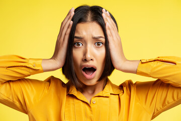 Portrait of serious, excited asian woman holding head with hands isolated on yellow background