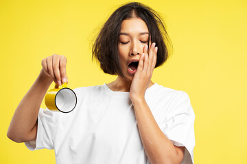 Portrait of tired young pensive Asian woman wearing white t shirt holding cup, yawning