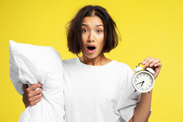 Portrait of shocked disheveled asian woman holding alarm clock and white pillow, late, overslept