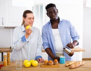 Happy biracial couple laughing and drinking coffee in morning in kitchen