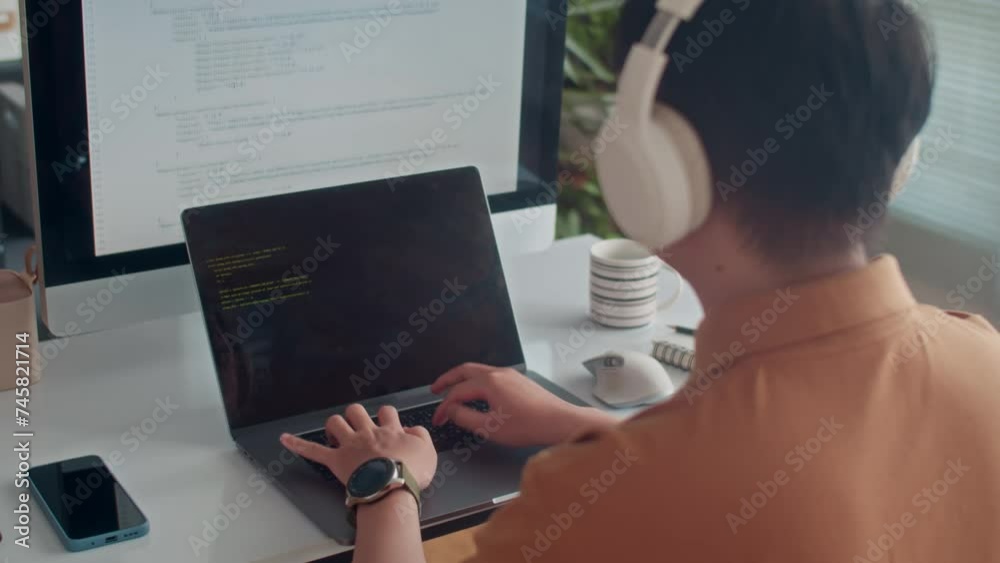 Wall mural back view of male programmer in headphones coding on laptop sitting in front of big monitor at offic