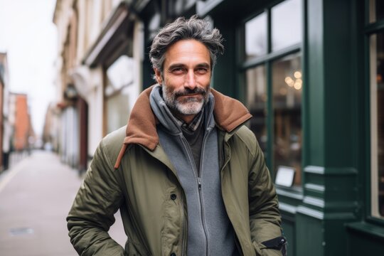 Portrait of a handsome bearded middle-aged man with gray hair in a green jacket and gray scarf on a city street.