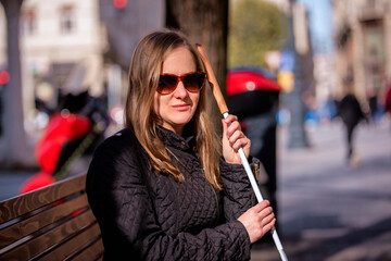 A visually impaired woman holding a white cane and sitting on a bench in the city