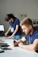 Young technician sitting by workplace in repair center office and fixing tiny details of broken mobile phone with screwdriver against colleague