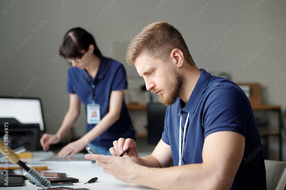 Poster young technician checking battery of broken mobile phone while sitting by workplace in repair center