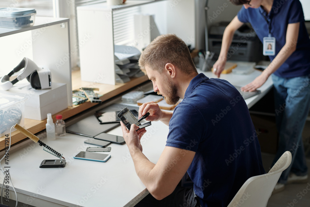 Poster young worker of technical repair office fixing tiny details of disassembled cellphone with small scr