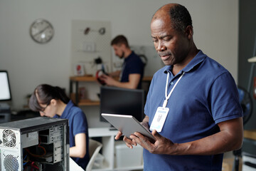 Serious male technician with tablet standing in front of camera in coworking space and looking through online instructions