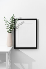 Black picture frame mockup on white wall with copy space, fresh eucalyptus twigs in vase decor