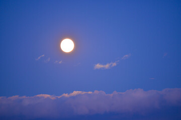 Full moon rising on the sky seen trough brightly clouds during summer season at dusk