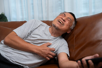 Senior male asian presses hand to his chest has a heart attack suffers from unbearable pain in the living room, lonely men try using the smartphone to call for Emergency help.