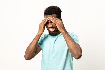 A young man in a blue t-shirt looks overwhelmed, covering his face with hands expressing stress, headache, or despair on a white background. - 745846179