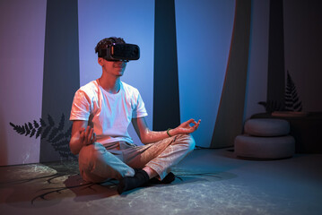 Smiling teenage programmer boy sitting on the floor with a virtual reality headset on, making hand gestures, typing, programming and scrolling in the air.