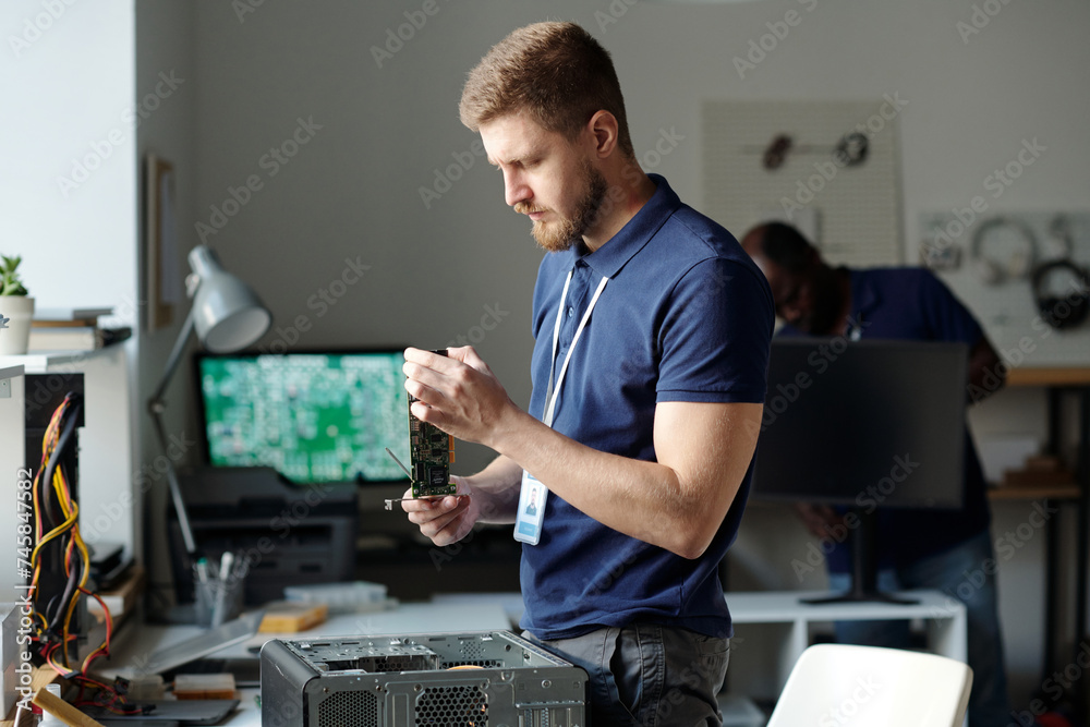 Poster Young male engineer of repair center checking detail of disassembled computer processor while standing by desk with hardware