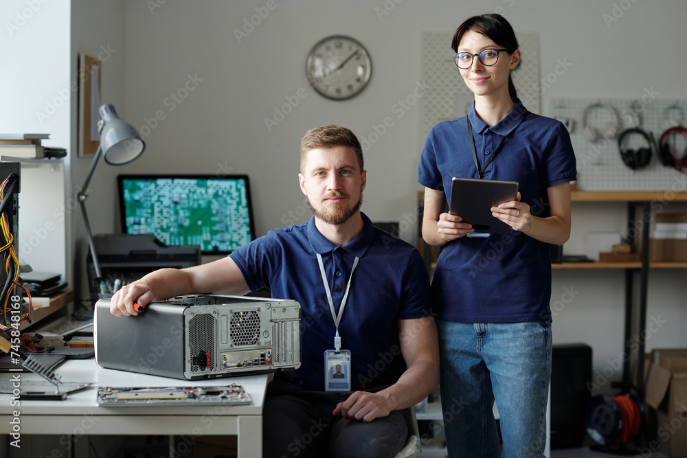 Poster team of two young successful engineers of repair service center in blue uniform looking at camera wh