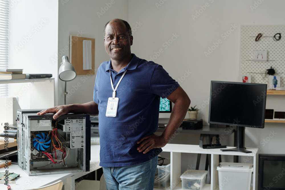 Poster Happy experienced engineer of maintenance center looking at camera with smile and keeping hand on top of computer processor in office