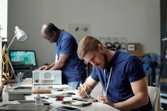 Young serious specialist of troubleshooting and repair service center with transmission tweezers bending over workplace with motherboard