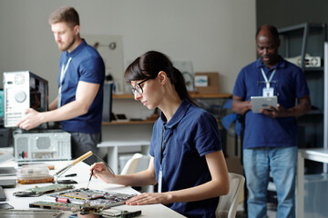 Obraz premium Young female technical service specialist fixing details of disassembled laptop with screwdriver against intercultural male colleagues