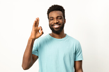 Cheerful young African male smiling and gesturing okay sign, signifying approval and positivity against a plain light backdrop. - 745850739