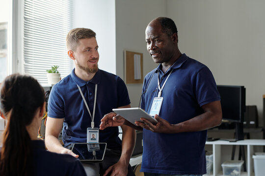 Confident mature businessman with tablet looking at one of young subordinates during explanation or discussion of working points