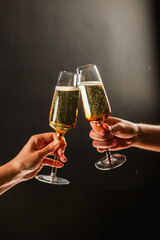 Two people clinking glasses of sparkling champagne or prosecco against a dark black background, with only their hands and the glasses visible. The moment captures a celebratory and atmospheric mood, h