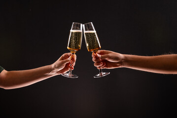 Two people clinking glasses of sparkling champagne or prosecco against a dark black background,...