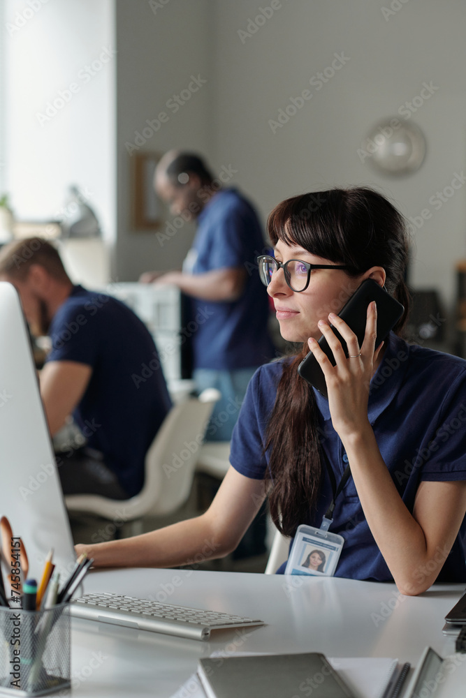 Poster young businesswoman in eyeglasses looking at computer screen and clicking mouse while talking to cli