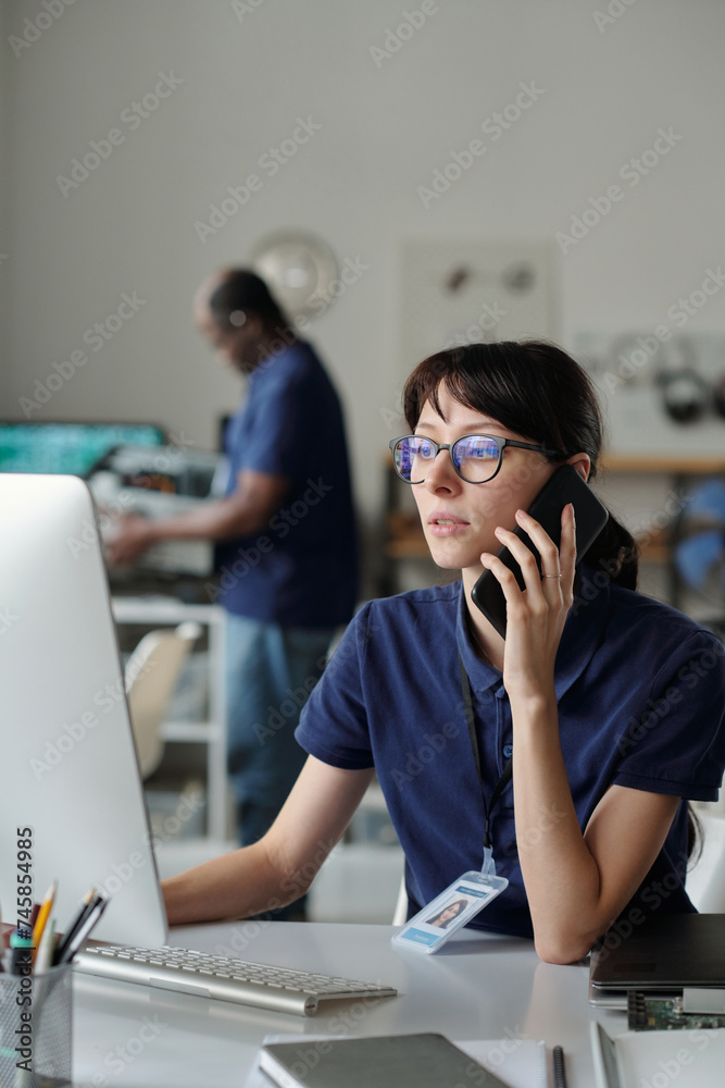 Poster Young confident technician with smartphone by ear communicating to client while sitting by workplace in front of computer screen