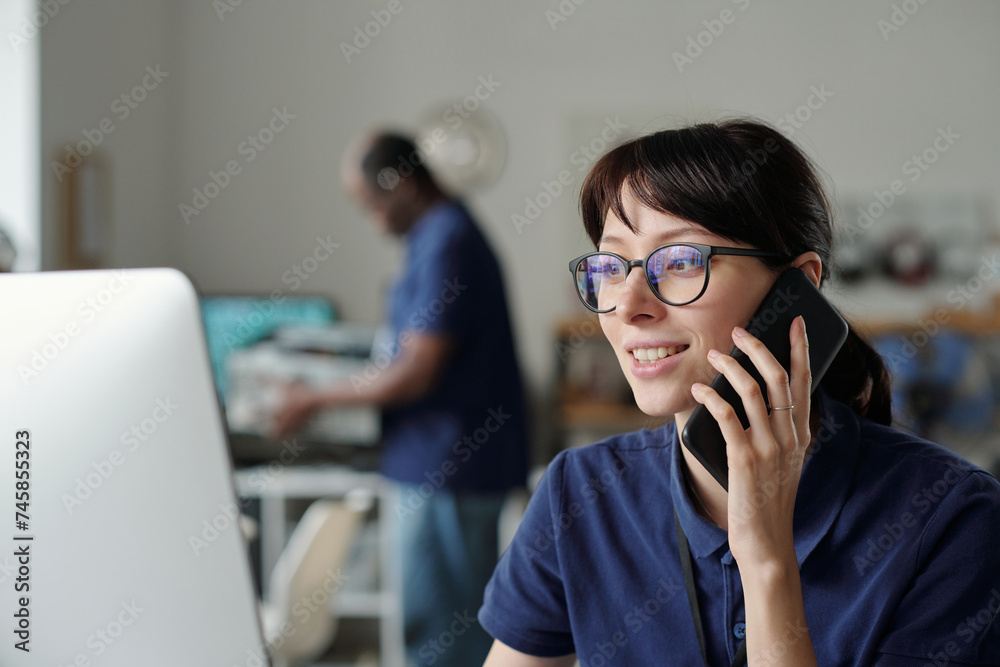 Poster Smiling female technician with mobile phone talking to client while sitting in front of computer screen and looking through online data