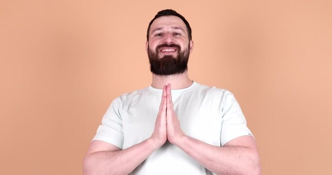 Portrait bearded man thank gesture with connected hands and reverence smiling posing isolated on beige background. Calmness happy male arms namaste praying yoga position with positive emotion.