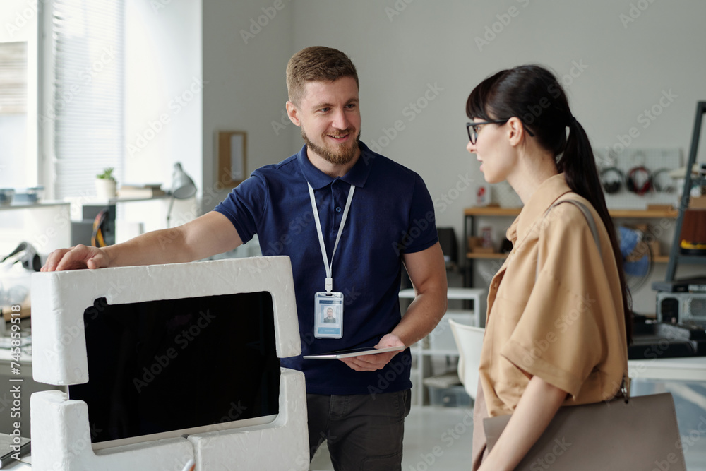 Poster Young smiling male manager of troubleshooting center keeping hand of packed computer monitor while giving it back to client after repair